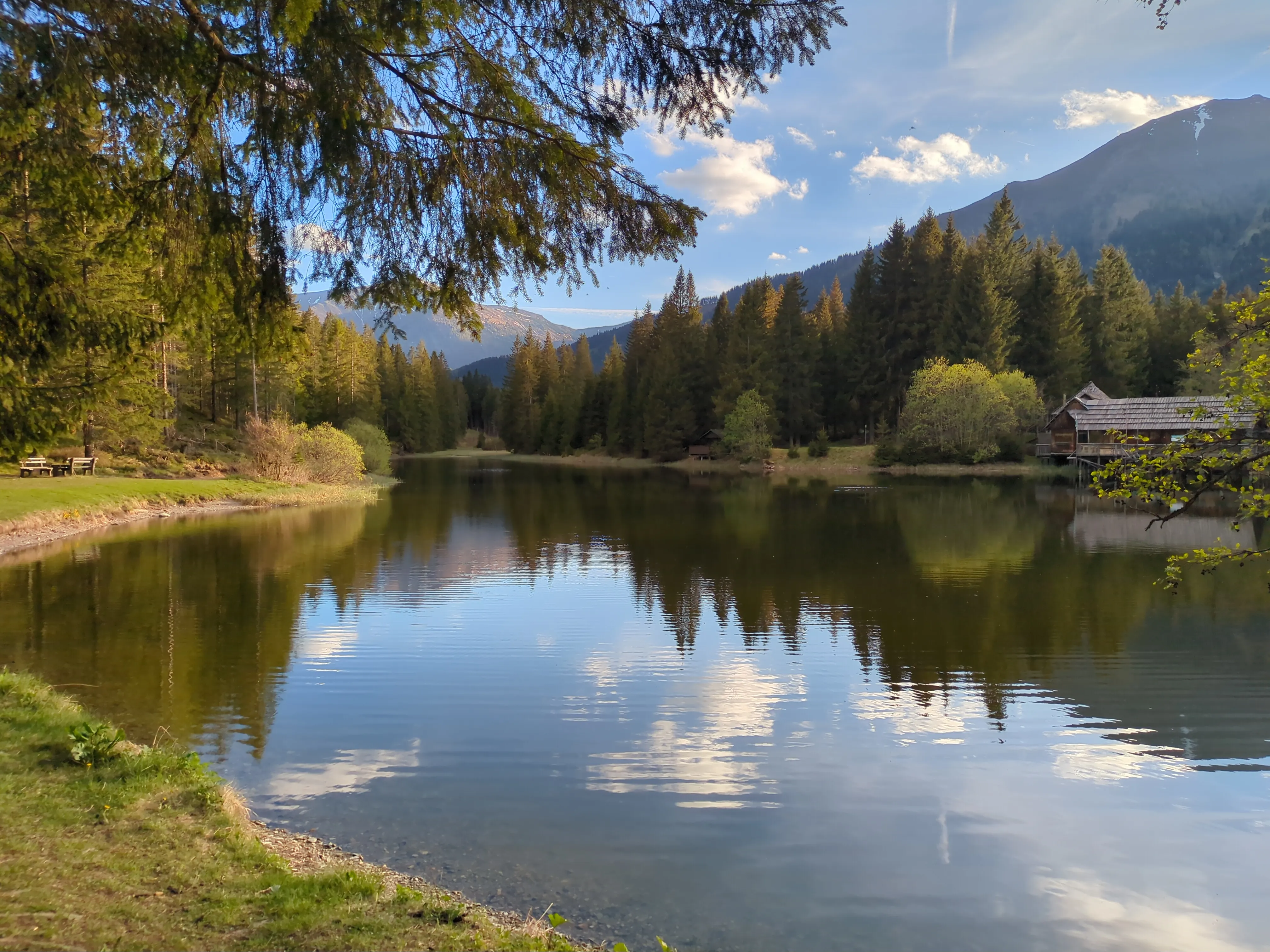 Ein natürlicher Teich, in dem sich die Wolken spiegeln, nimmt den Großteil des Bildes ein. Rechts im Hintergrund liegt das keltische Pfahlbaudorf. Grüne Wiesen und Bäume. Eine Abendstimmung, hervorgerufen durch den einsetzenden Sonnenuntergang.